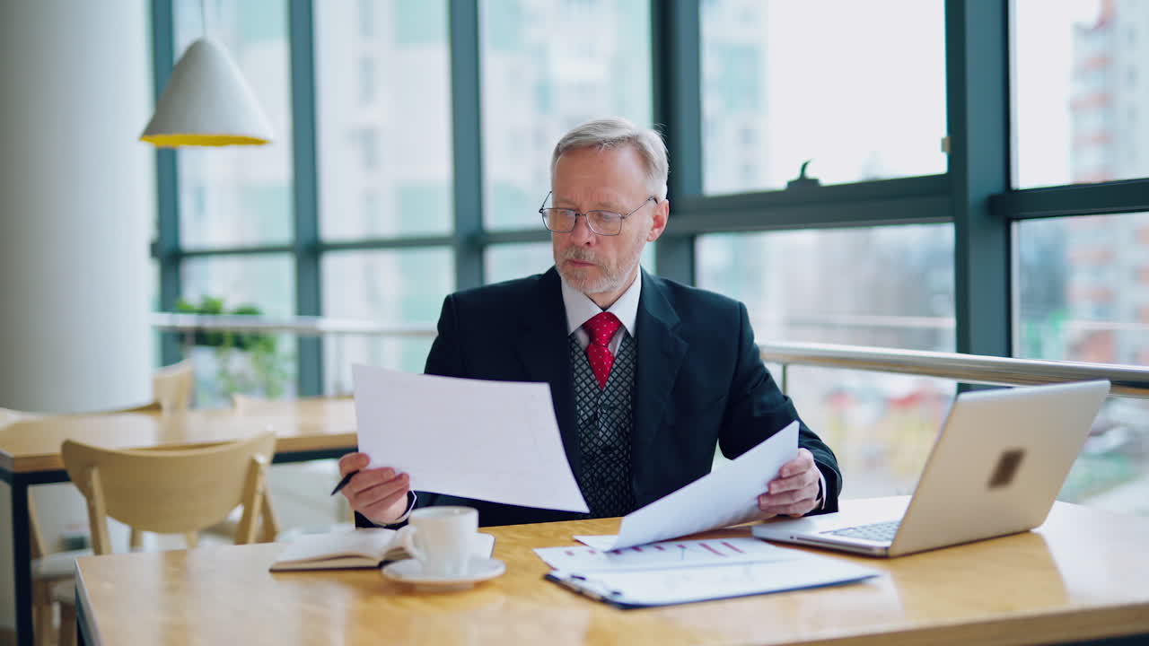 Businessman is reading a printed report or document while sitting at the table, working on paper work with laptop at the table. Man looking at documents. Business video.