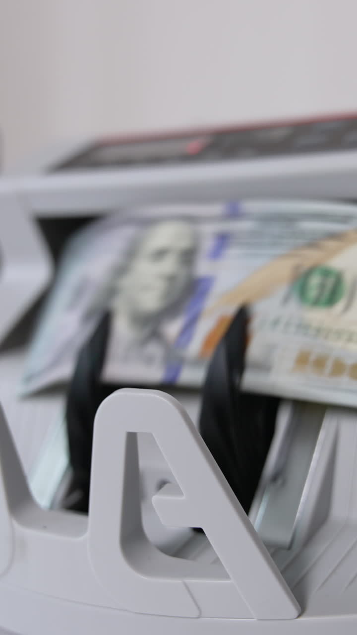 A hand switches on money counting machine. A wad of hundred dollar bills is being put into equipment for cash calculation. Close up. Blurred backdrop. Vertical video