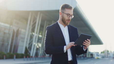 Portrait-Of-The-Good-Looking-Young-Businessman-Taping-And-Scrolling-On-The-Tablet-Device,-Then-Turning-His-Head-And-Smiling-To-The-Camera-Near-The-Airport-Or-Train-Station