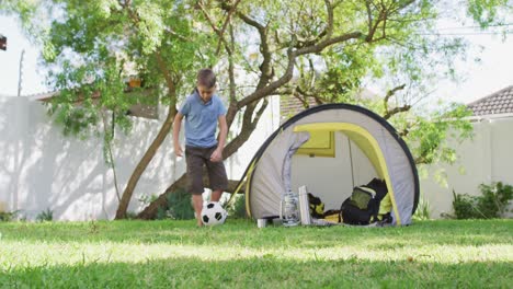 caucasian boy playing with football in the garden on a sunny day by a tent