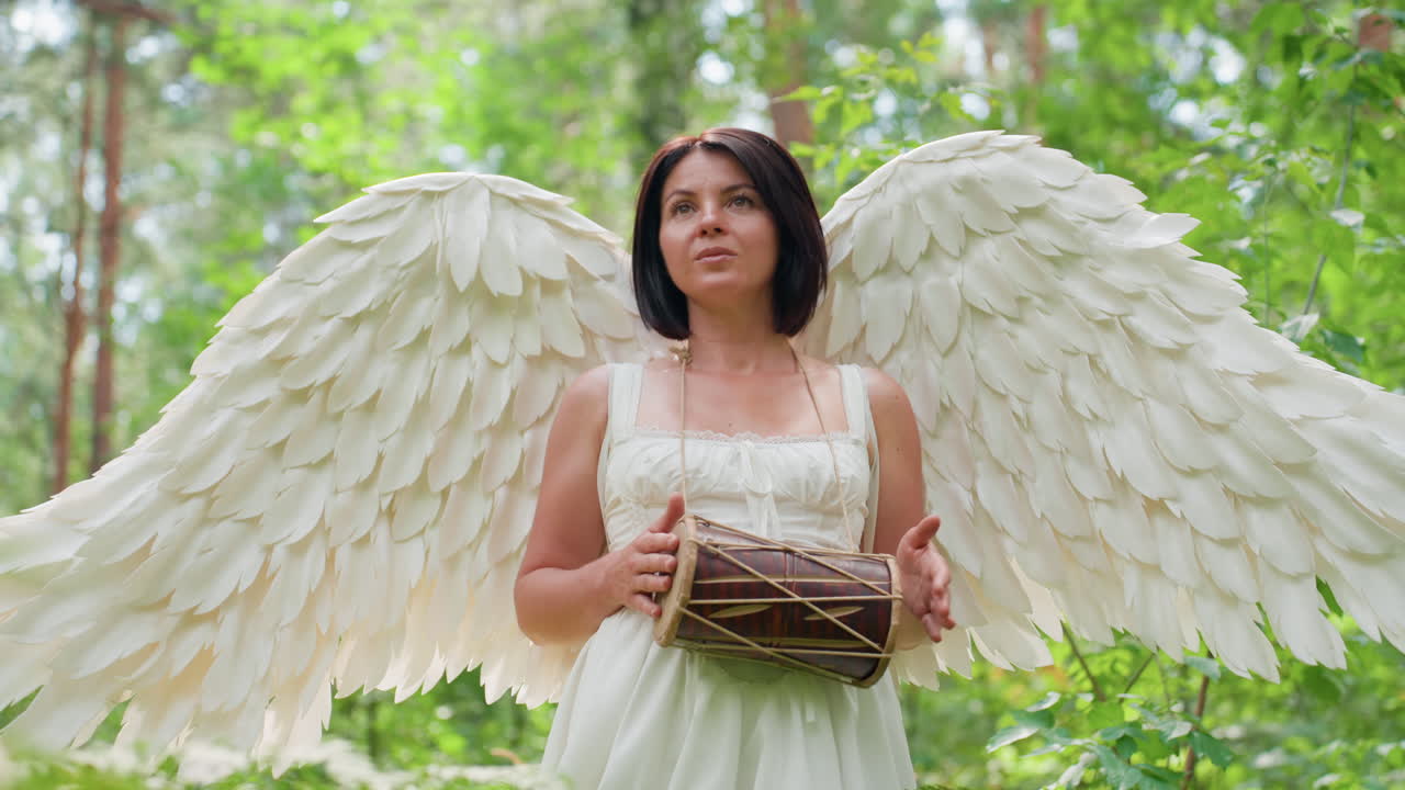 Close up of fairy woman in white dress with wings walking in green forest while beating hand drum, soft sunlight on hands and leather skin, gentle rhythm, cultural vibe, peaceful nature mood