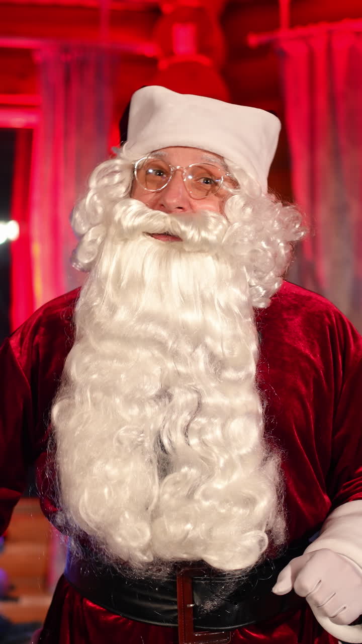 Portrait of Santa Claus indoors. Aged man in Santa Claus costume with natural beard congratulations with Christmas. Elderly man talking on camera wishing merry Christmas. Vertical video