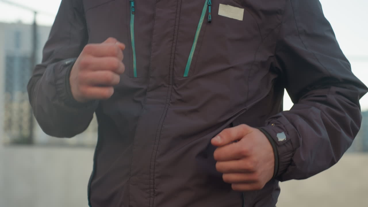 primer plano de un hombre corriendo al aire libre con chaqueta, mostrando el movimiento del brazo y el equipo de fitness, enfatizando el movimiento dinámico, la determinación y el estilo de vida activo contra un telón de fondo urbano borroso