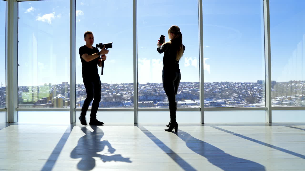 Photographer and a model with a phone indoors. Young woman doing selfie photos on her phone while man taking photos of woman on a professional camera.