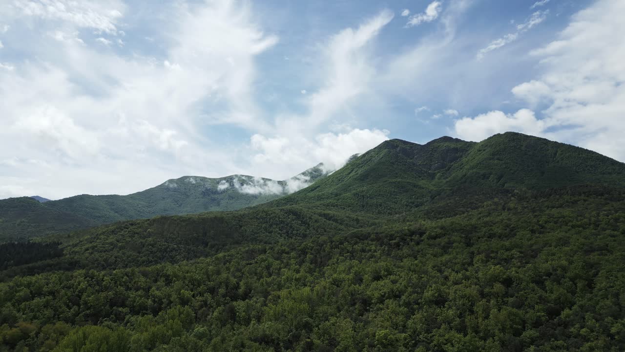 Majestic mountain peaks rising towards blue sky, covered in lush forests, with ethereal clouds drifting around summits, creating breathtaking vista of pristine wilderness