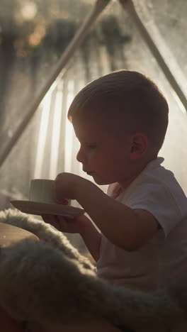 kid with tea cup at small table at glamping. calm boy takes sip of brewed drink on floor in evening camp. child enjoys beverage at night camp