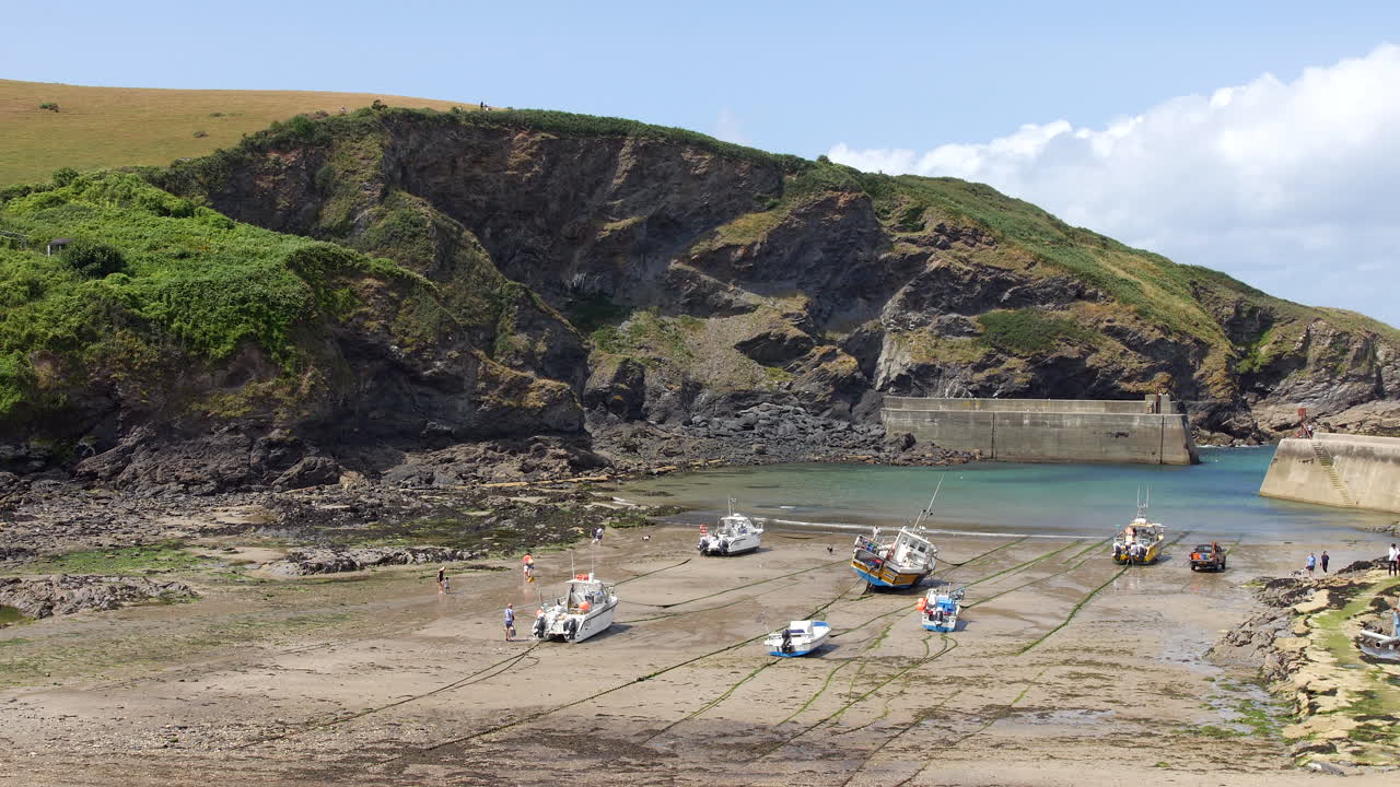 Boats lying on wet sand during low tide are tethered across a harbor surrounded by cliffs and breakwaters, as people walk between exposed rocks and shallow pools Port Isaac, Cornwall, England