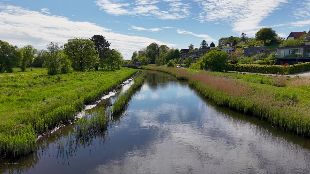 Drone video captures a calm canal lined with green grass trees and houses reflecting a blue sky with clouds in rural Denmark on a sunny day