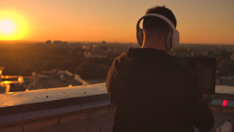 A-man-in-headphones-on-the-roof-relaxes-working-remotely-enjoying-life-despite-a-handsome-kind-of-sipping-beer-and-types-on-the-keyboard.-Trade-on-the-stock-exchange-using-a-laptop-and-enjoying-the-beautiful-view