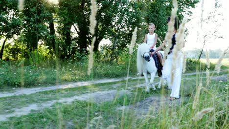 a woman and a boy are walking around the field, son is riding a pony, mother is holding a pony for a bridle. cheerful, happy family vacation. outdoors, in summer, near the forest