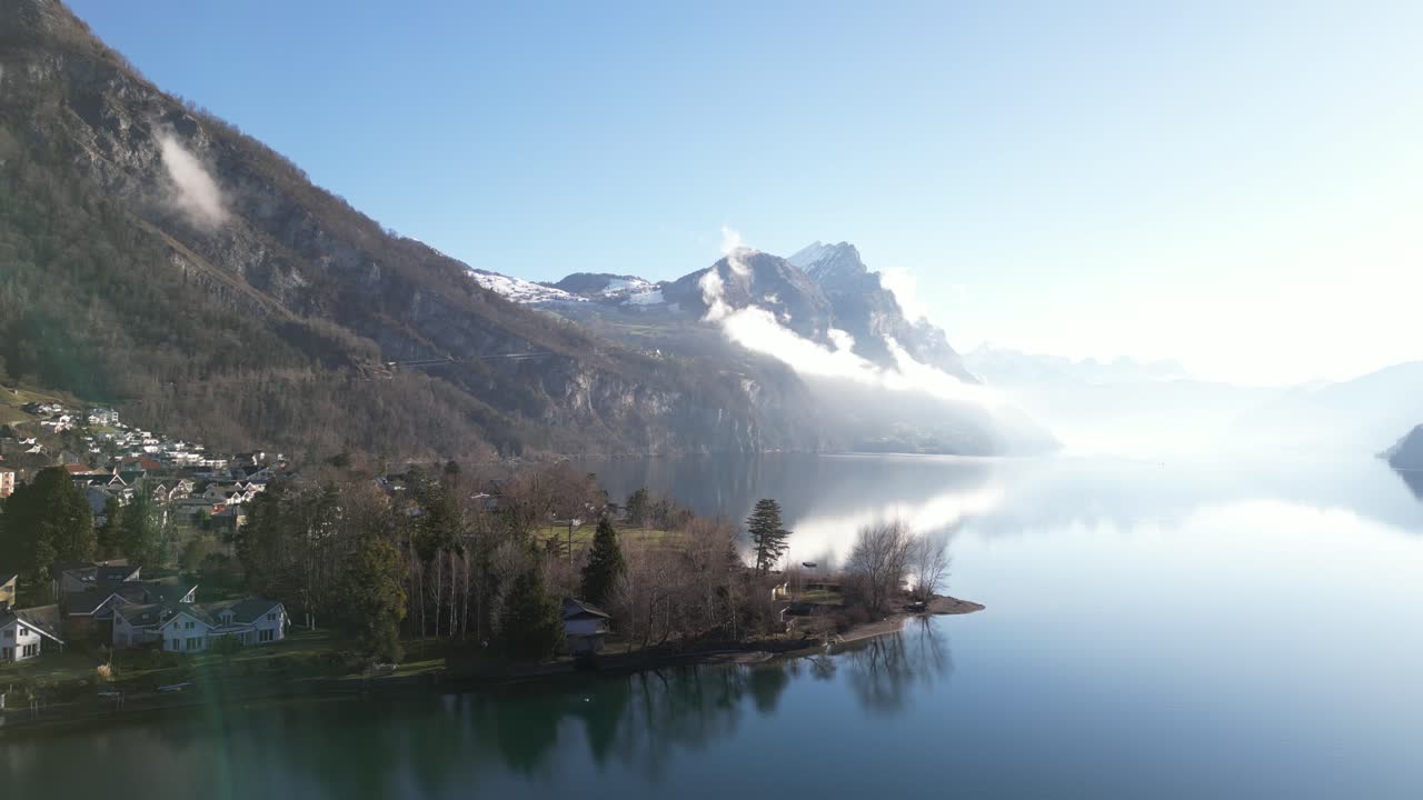 un clip brillante de un dron con una llamarada de lente que muestra montañas cubiertas de nieve con niebla junto al lago calm en suiza