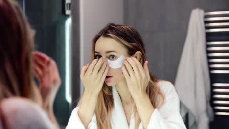 mujer joven en bata de baño aplicando parches blancos en los ojos mientras mira al espejo