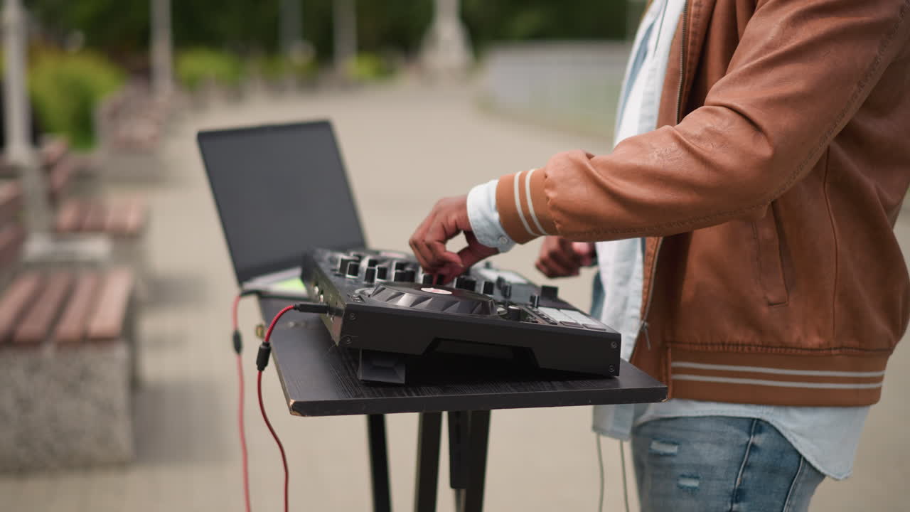 DJ:n kädet mikserillä ulkopuistossa lähikuvassa, sormet säätävät nuppeja, kannettava tietokone ohjaimen vieressä, punainen johto roikkuu, rento takki ja farkut, penkkien reunustama kävelytie, päiväaikaan keskittynyt taktiilinen tunnelma, katu