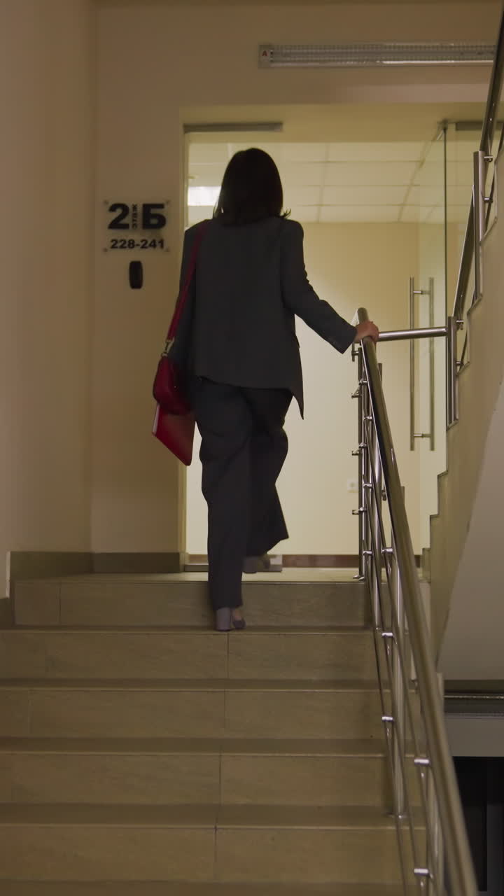 Businesswoman walking upstairs in office building carrying red leather handbag, focused and confident. Stylish and professional in office attire, heading to work with determination and focus