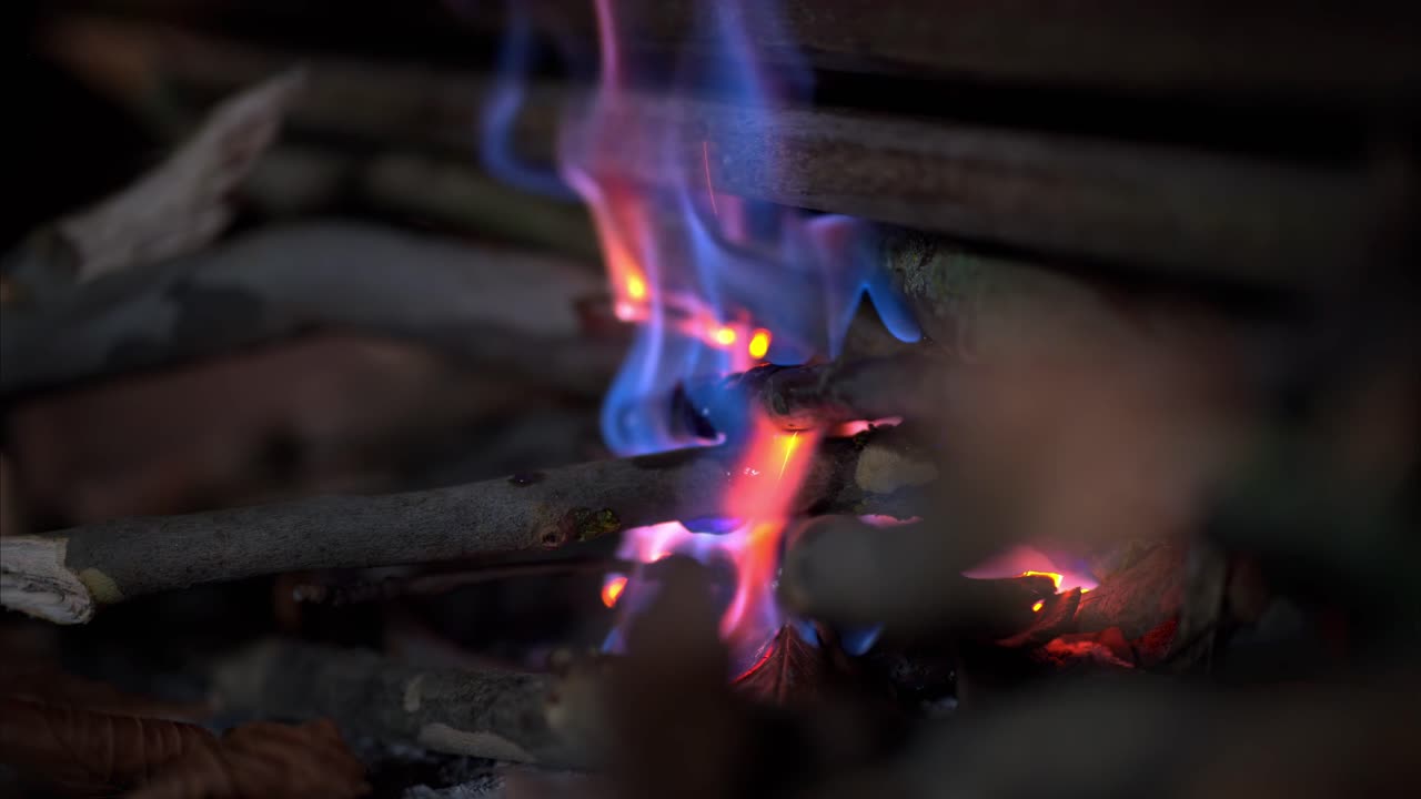 Close-up de gravetos de madeira queimando com chamas azuis e laranjas, brasas brilhantes e luz suave de fogueira tremeluzindo em um ambiente interno escuro