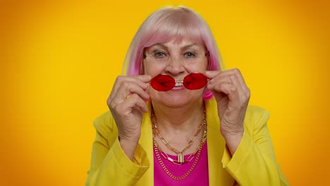 una mujer mayor con cabello rosa sonríe mientras lleva una chaqueta amarilla y gafas rojas