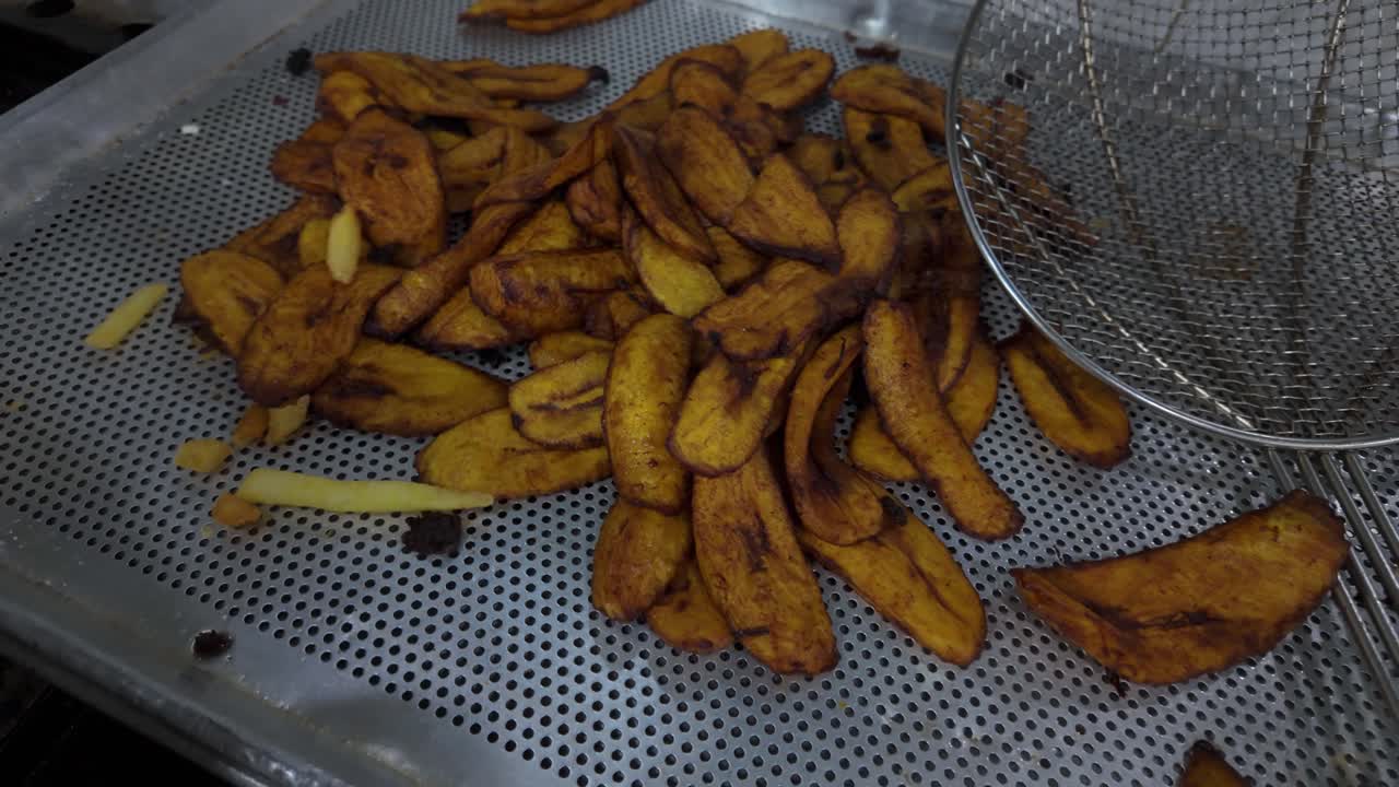 Vast pile of sweet, golden-brown fried ripe plantain slices (tajadas) cooling on a perforated stainless steel tray in commercial kitchen, with metal skimmer nearby