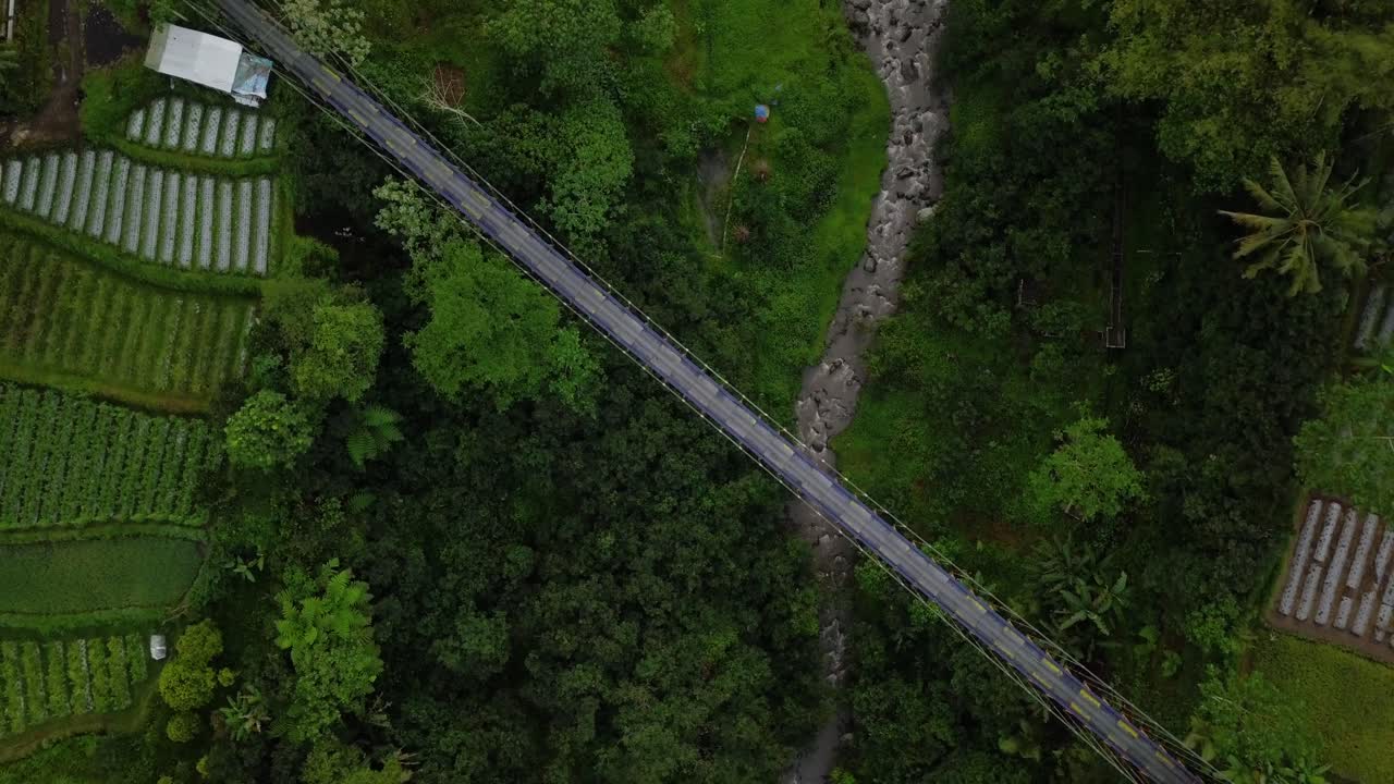 ภาพโดรนแนวตั้งของสะพานแขวนโลหะที่สร้างพาดผ่านหุบเขา โดยมีแม่น้ำอยู่ด้านล่าง และล้อมรอบด้วยต้นไม้และไร่ผัก