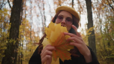 smiling lady in a yellow beret and black coat adjusting a vibrant autumn leaf, gazing upward with a joyful expression in a sunlit forest