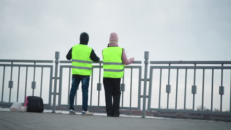 two people in reflective safety vests standing near metal fence by waterfront on cloudy day, holding bottled water, observing scenery, backpacks on ground, overcast sky, outdoor work break