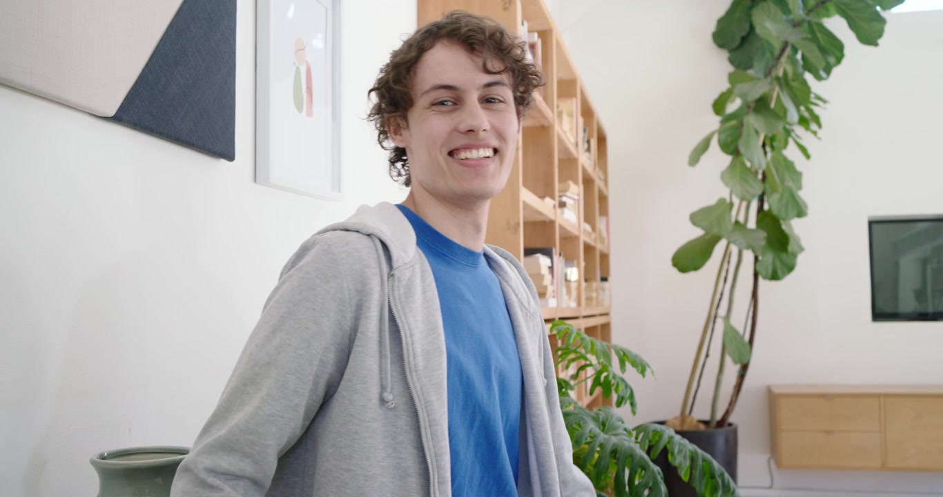 Uomo con maglietta blu tiene il telefono, scorre lo schermo, sorride a un messaggio e si gira verso la fotocamera in soggiorno