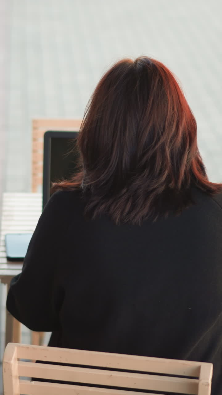 vista trasera de una mujer trabajando en una computadora portátil al aire libre en una mesa de madera con un teléfono inteligente a su lado, rodeada de suelo pavimentado y una vista parcial de flores