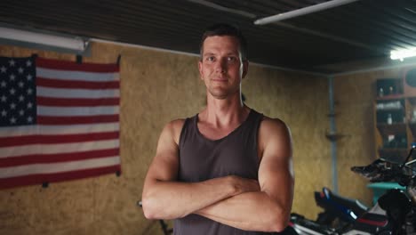 Close-up-portrait:-confident-mechanic---motorcyclist-folds-his-arms-across-his-chest-and-looks-at-the-camera-in-his-workshop-against-of-the-US-flag