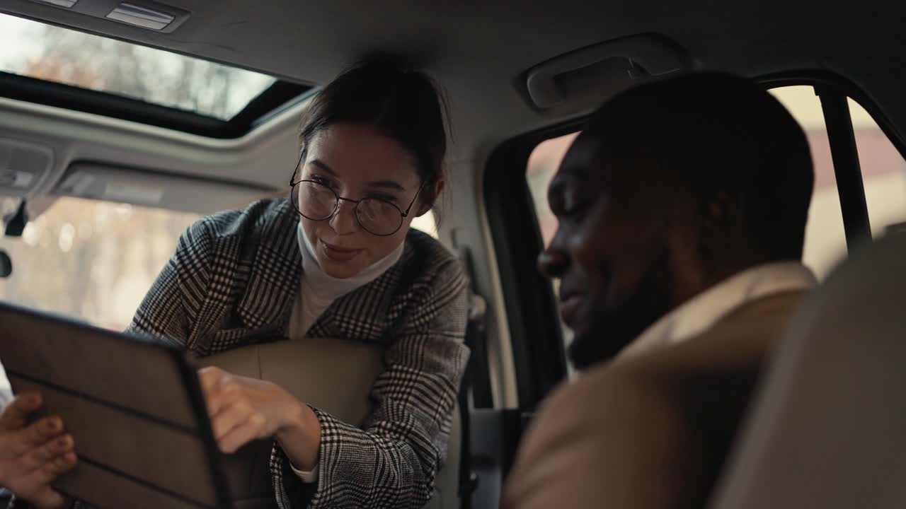 A happy and confident brunette girl in round glasses in a suit communicates with a Black-skinned businessman in a brown jacket and presents her plans using a tablet during her car ride and business trip