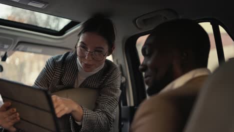 a happy and confident brunette girl in round glasses in a suit communicates with a black-skinned businessman in a brown jacket and presents her plans using a tablet during her car ride and business trip