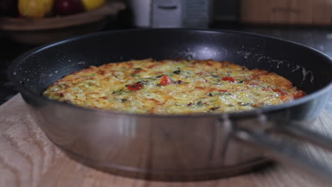 Slow-Pan-right-to-left-of-cooked-golden-brown-tomato,-red-pepper,-kale,-zucchini--courgette,-bacon-and-onion-omelette-frittata-in-frying-pan-on-wooden-chopping-board