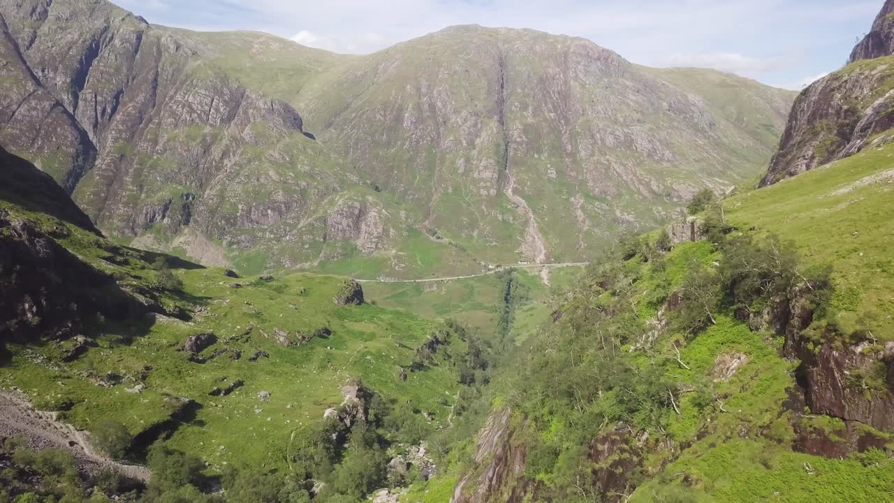 Prato verde in primo piano con le Tre Sorelle Montagne sullo sfondo e una gigantesca valle sottostante, Glencoe Scozia in una giornata parzialmente nuvolosa