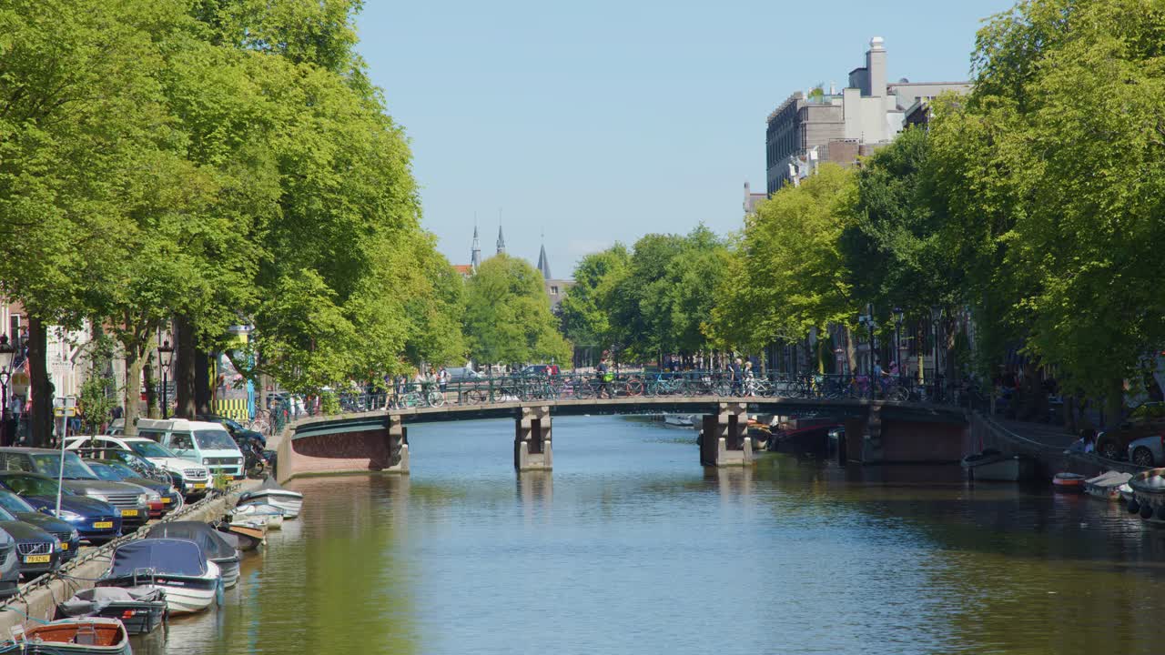 Wide shot of Amsterdam canal, bridge, cyclists, and boats under bright daylight, static camera