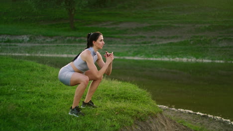 training in the park of a woman performing squats and jumps on the background of the river in the park. do sports in the morning near the lake