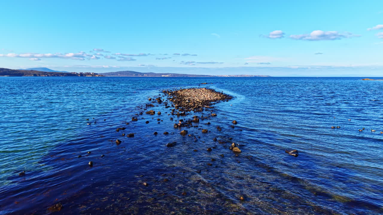 Serene rocky path leading into a clear blue sea under a vibrant sky filled with soft clouds at midday