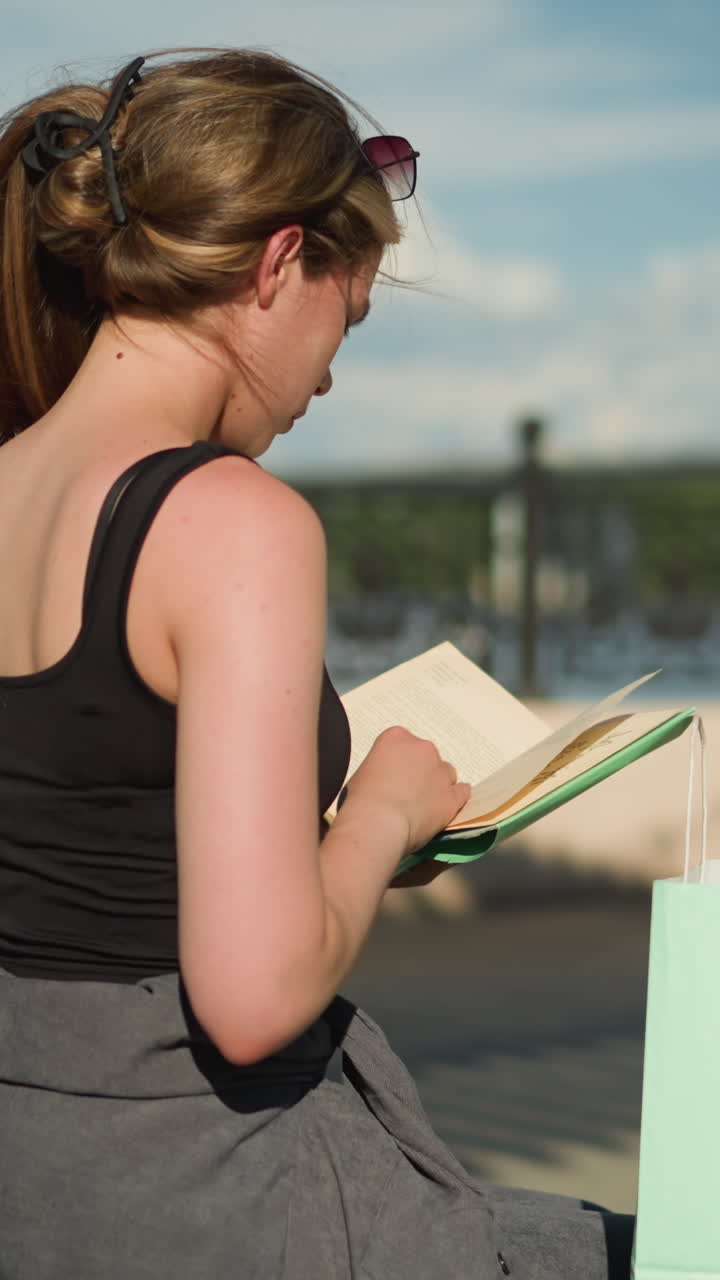vista trasera de una mujer sentada en un banco al aire libre con bolsas de compras a ambos lados, ella recupera un libro de una bolsa verde menta, lo abre y hojea sus páginas