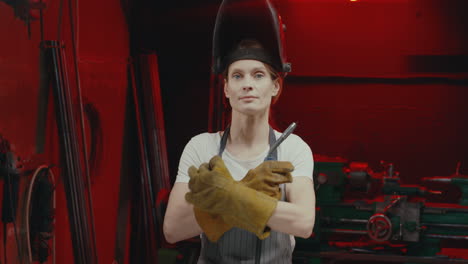 caucasian young woman welder taking off metal protective mask and looking at camera with arms crossed in workshop