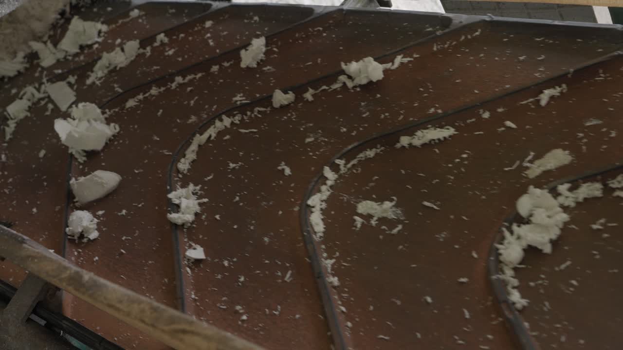Primo piano di scaglie e frammenti di plastica bianca sparsi su un nastro trasportatore in movimento in un impianto di riciclaggio.