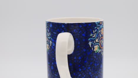 Decorative ceramic mug featuring intricate floral designs on a deep blue background, slowly rotating to showcase its detailed artwork
