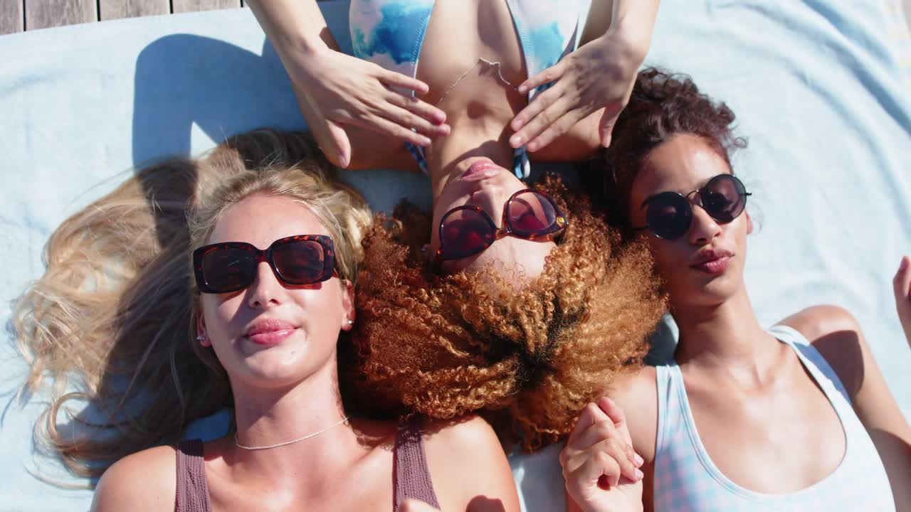 Diverse women friends lying on deck towel in swimwear sunlight making center pal adjusting shades