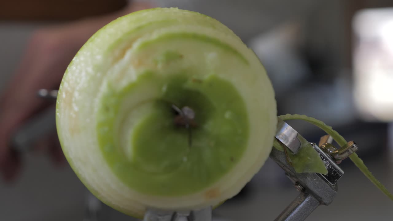 Visione frontale al rallentatore di una mela verde che viene girata e sbucciata da un dispositivo da cucina per rivelare la polpa sul bancone, con il succo che schizza via e bassa profondità di campo.