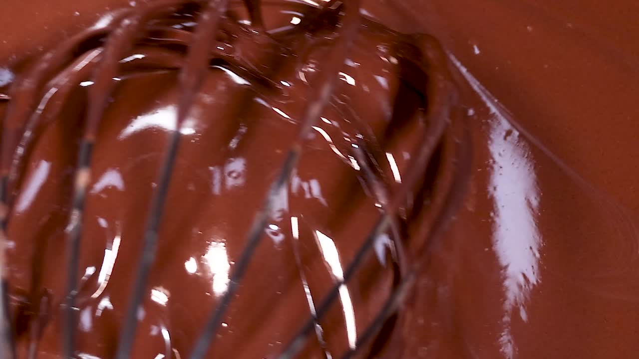 A whisk gently stirs through smooth, glossy melted chocolate, creating rich textures and reflections.