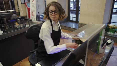 Cheerful-slender-saleswoman-in-white-shirt-and-black-apron-scanning-product,-fruits-at-checkout-counter-in-bright-supermarket-and-putting-it-into-brown-paper-bag.-High-angle-footage