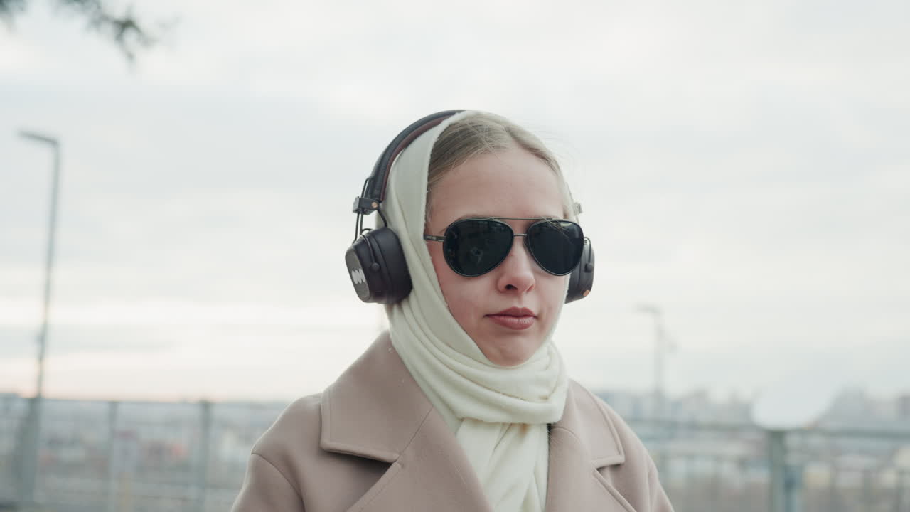 音楽を楽しむ女性。冬の装いでサングラスとヘッドホンを着用し、屋上で音楽のひとときを満喫する白人女性。