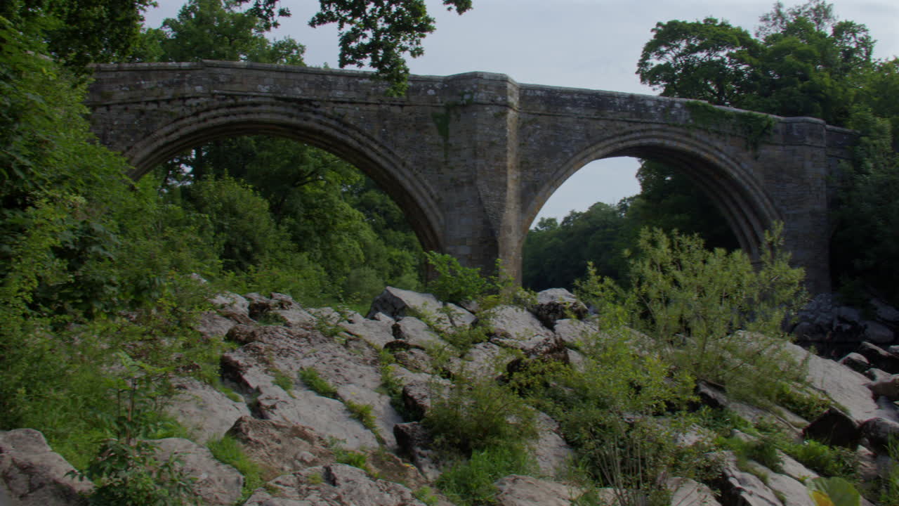 Wide panning shot looking up to devils bridge from the river Lune with rocks in foreground at Kirkby Lonsdale, North Yorkshire