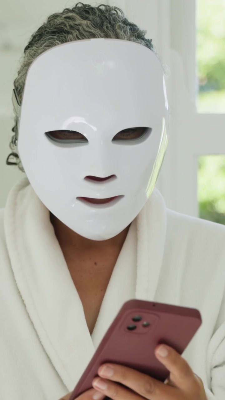 Vertikales Video: Frau im Bademantel hebt LED-Maske an, passt sie am Waschbecken an, tippt auf ihr Telefon und startet eine Sitzung.