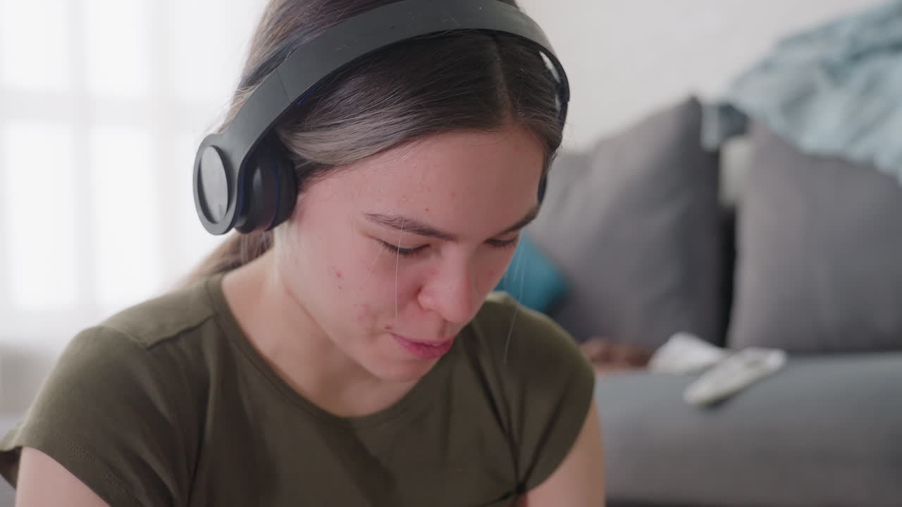 Close up of young woman wearing headphones enjoying music with relaxed smile, softly singing along with expression of joy, seated indoors in casual outfit with natural daylight