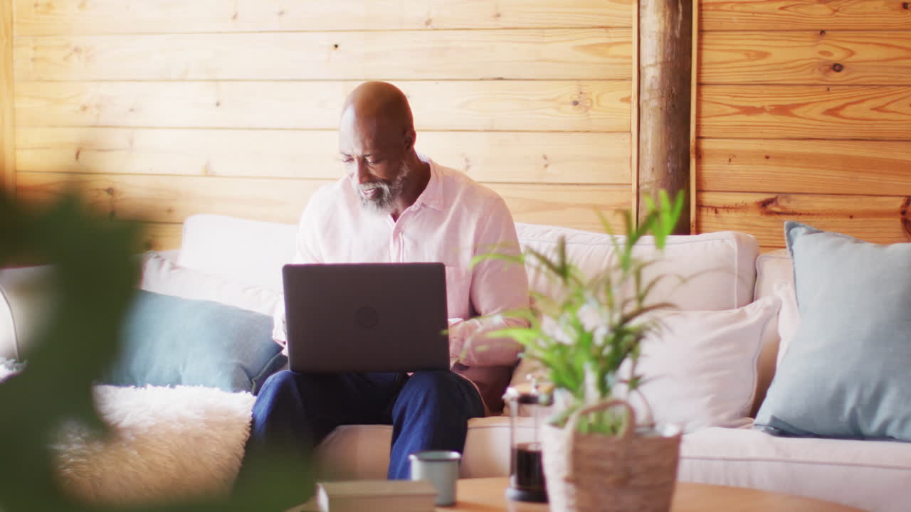 Happy senior african american man in log cabin, using laptop for online shopping, slow motion