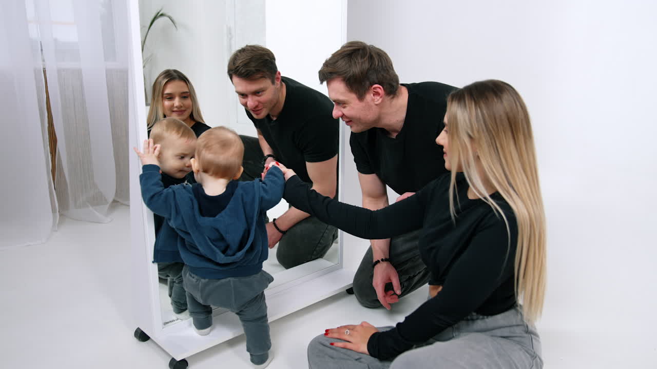 Little infant in blue sweatshirt and grey jeans stands at the mirror. Mother holds a baby by the hand and dad talks to his child.