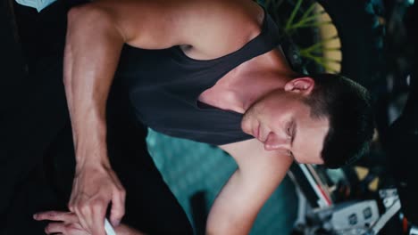 Vertical-video:-Close-up-shot:-a-male-mechanic-with-a-short-haircut-in-a-gray-shirt-sits-on-the-floor-and-looks-at-the-screen-of-his-white-smartphone-in-a-workshop-garage