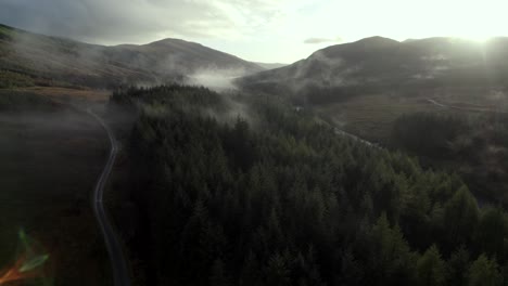 Drohnenaufnahmen-Aus-Der-Luft,-Die-Langsam-In-Einen-Dunklen-Nadelwald,-Eine-Straße-Und-Einen-Fluss-Hinabsteigen,-Während-Lichtstrahlen-Tief-Hängende-Wolken-In-Den-Baumwipfeln-Bei-Sonnenuntergang-Beleuchten